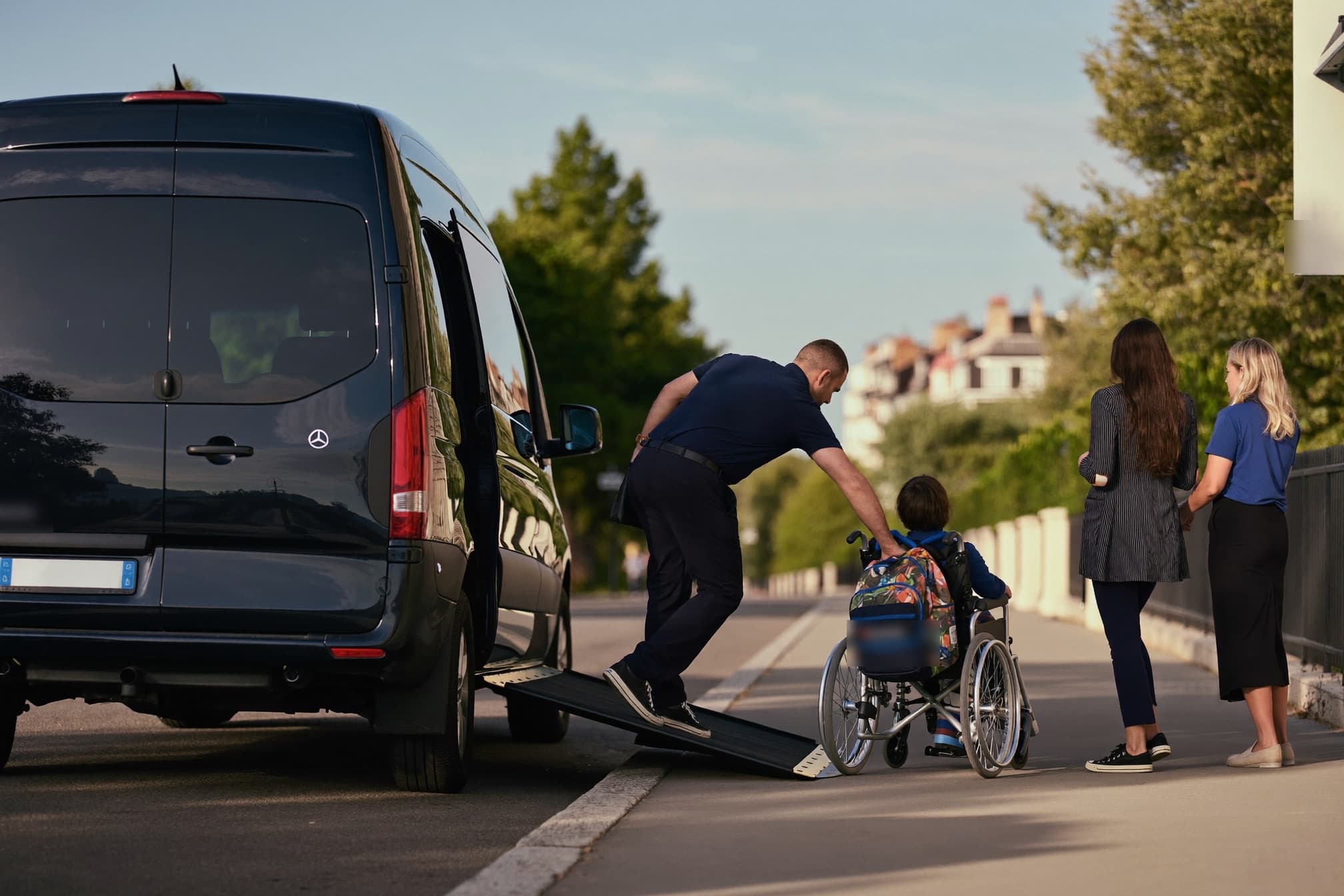 Un chauffeur déploie la rampe d'un véhicule adapté pour qu'un enfant en fauteuil roulant monte à bord, sous le regard de sa famille.